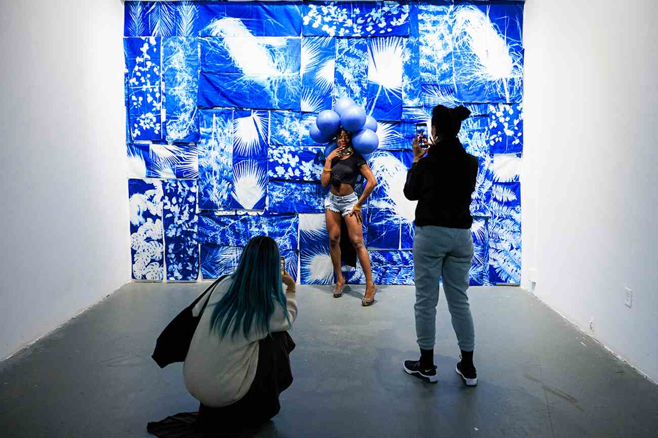 A person poses with balloons on their head, standing against a wall mural in blue and white, while two other people take pictures of them.
